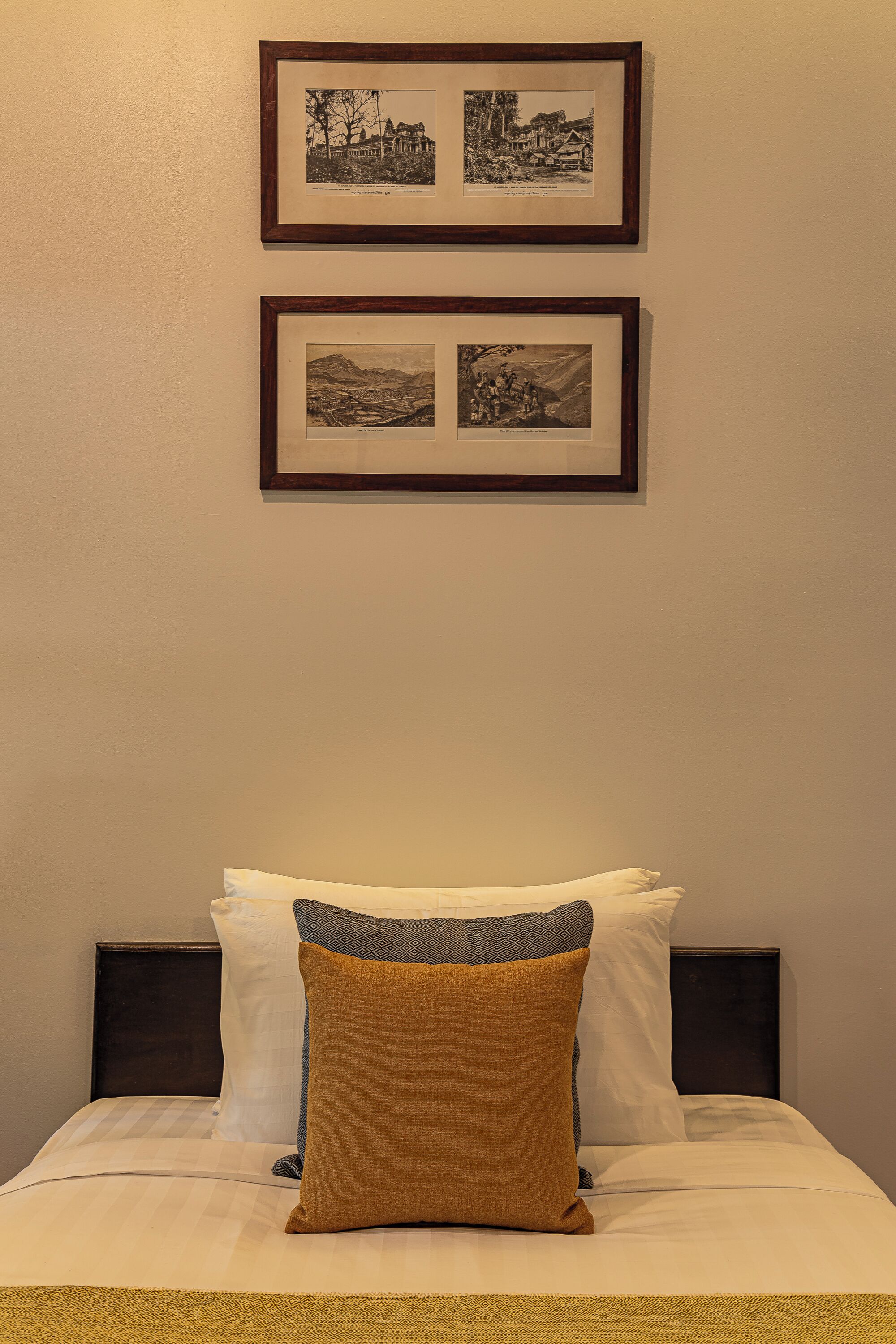 A neatly made bed with white and brown pillows, a framed picture above it, and a plain wall.