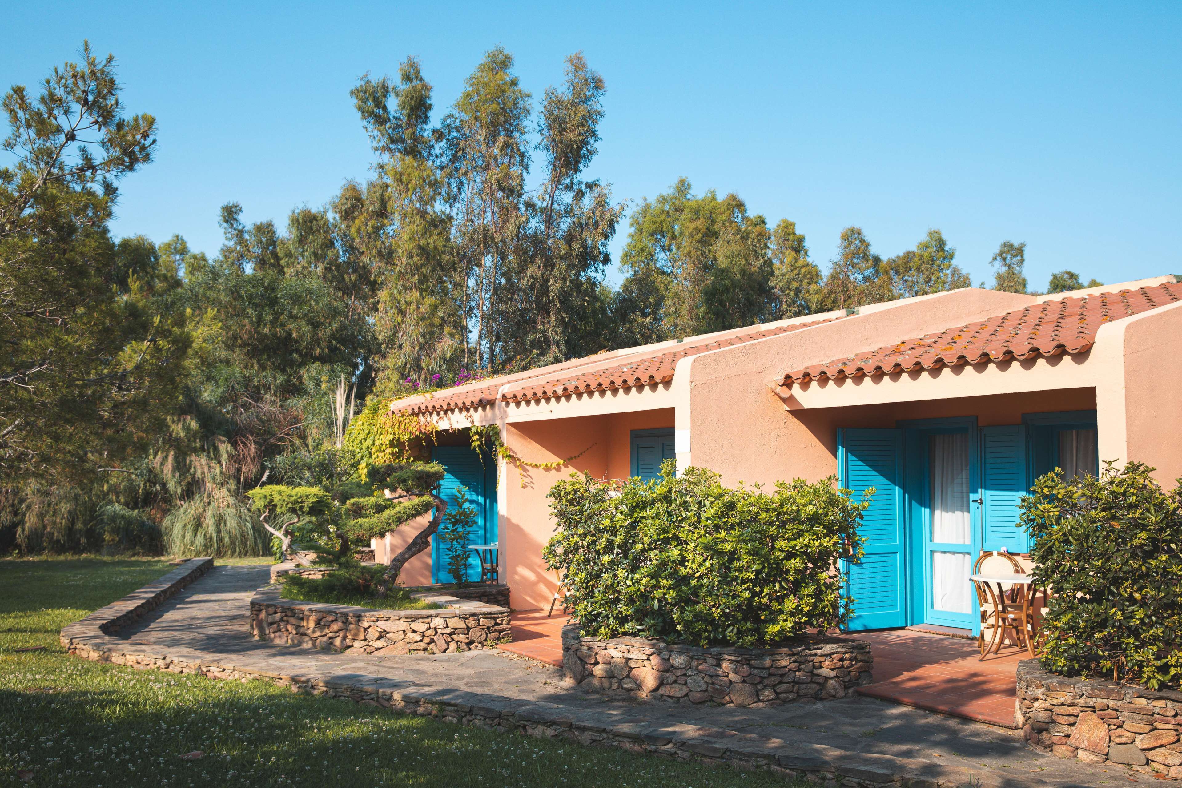 Un edificio de dos plantas con techo de tejas y puertas azules. Un camino de piedra conduce a la entrada, y el área circundante está llena de vegetación y árboles.