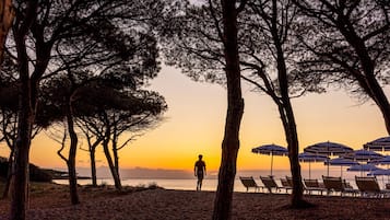 Une scène de plage où l’on voit une personne marchant vers le coucher du soleil, entourée d’arbres et de parasols de plage.
