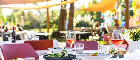 An outdoor dining area with tables set for two, featuring a variety of dishes and glasses of red wine.