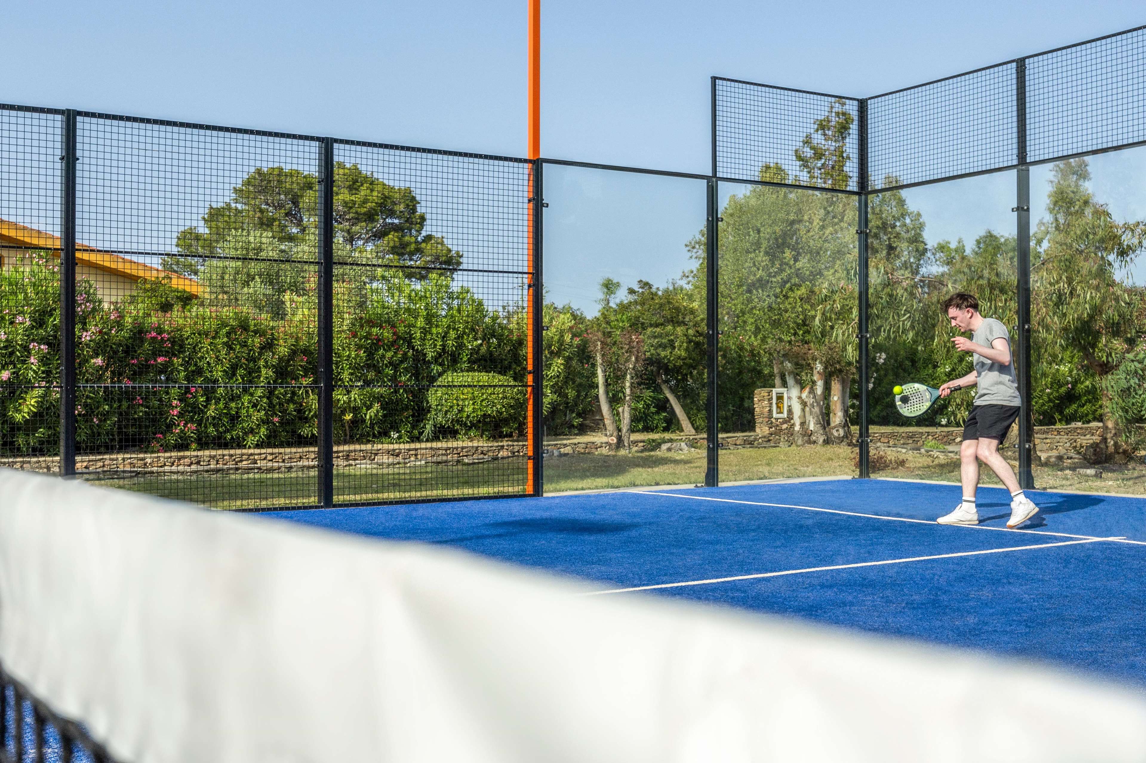 Ein Tennisplatz mit Netz, umgeben von einem schwarzen Zaun und Bäumen.