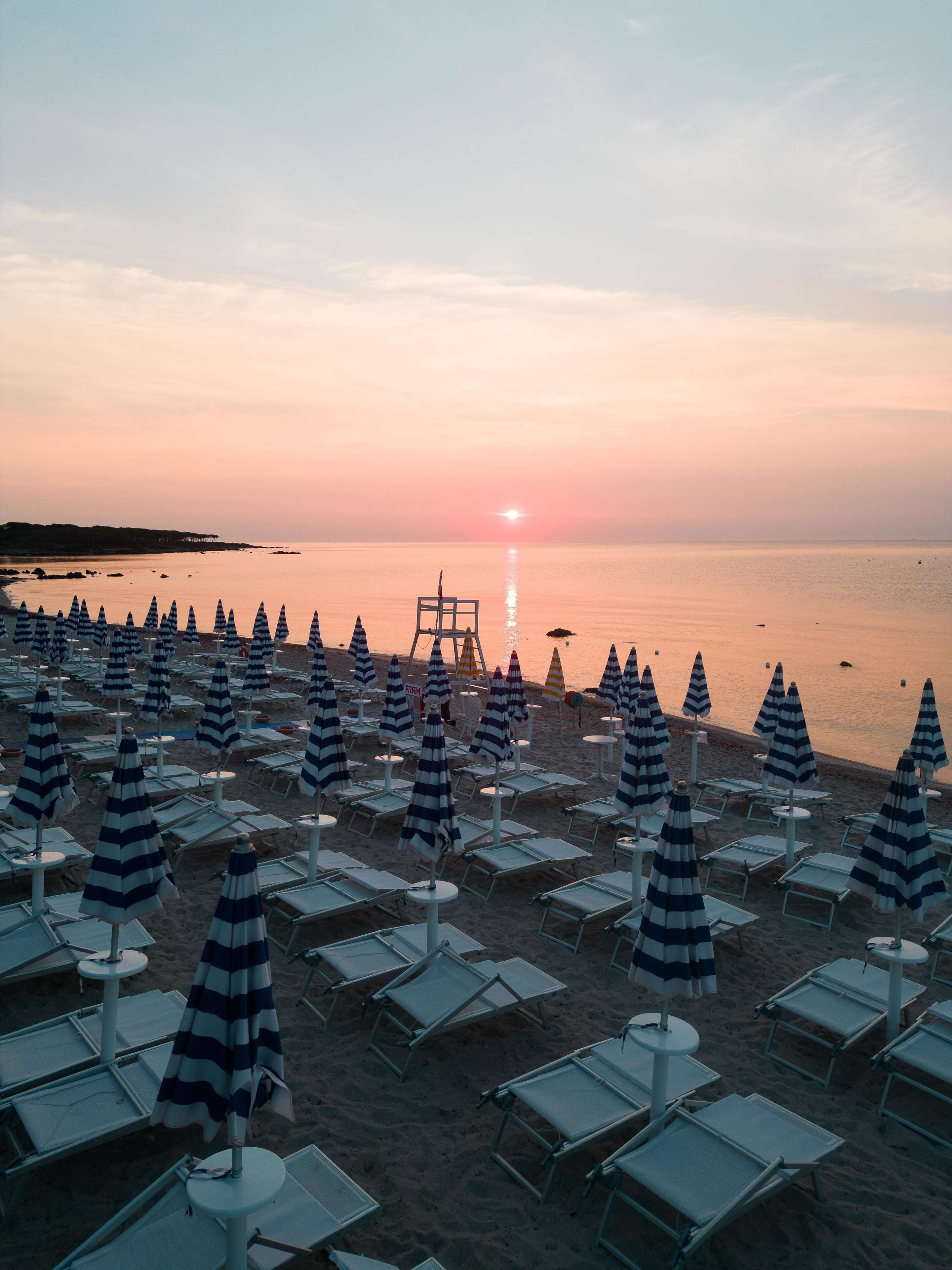A beach with rows of striped umbrellas and lounge chairs, a lifeguard tower, and a sunset over the water.