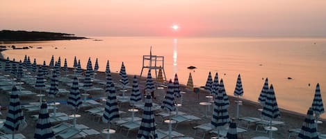 Una playa con hileras de sombrillas a rayas y sillas reclinables, una torre de salvavidas y un atardecer sobre el agua.
