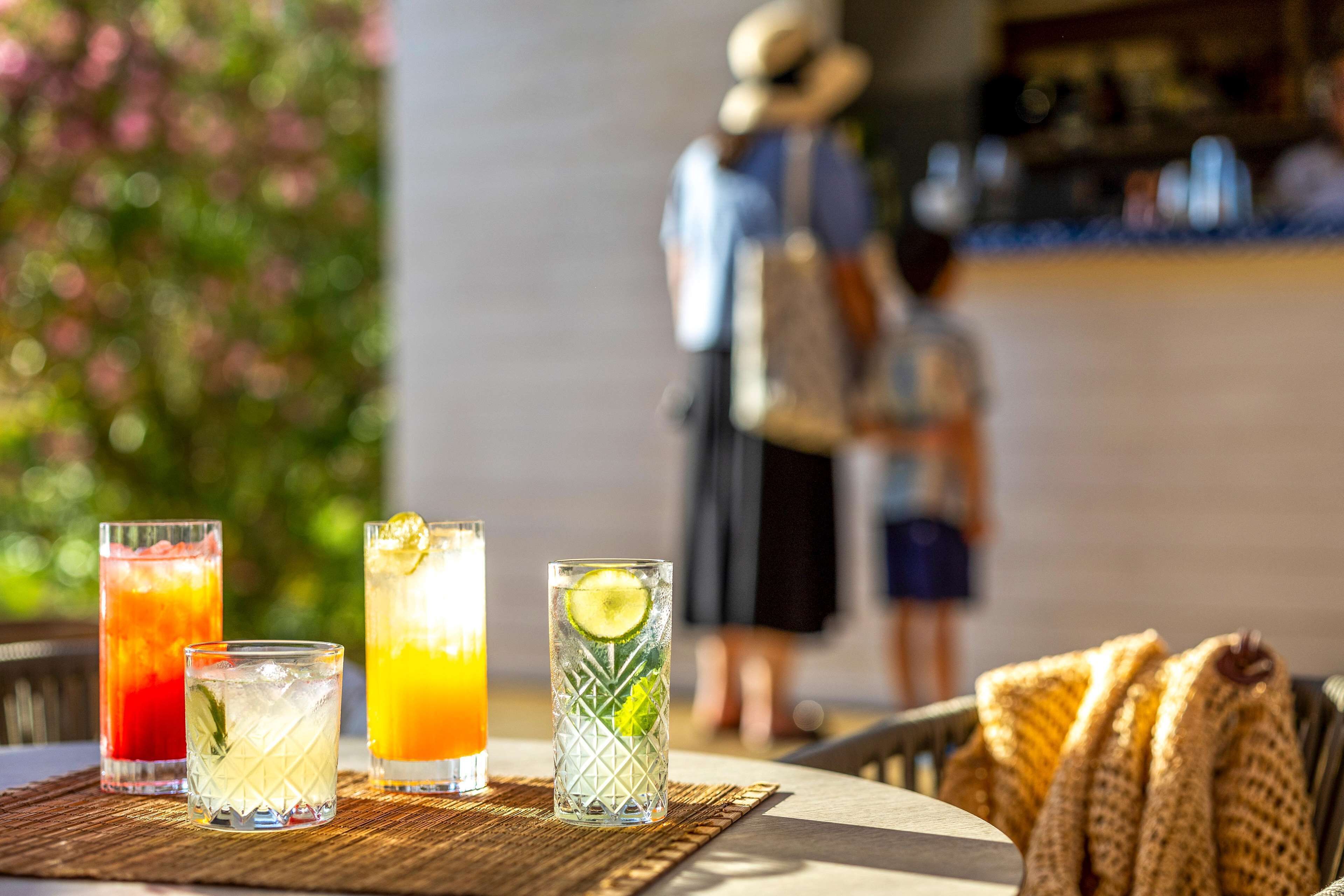 Three drinks on a table with a woven placemat, a basket of folded towels, and a blurred background of people and greenery.
