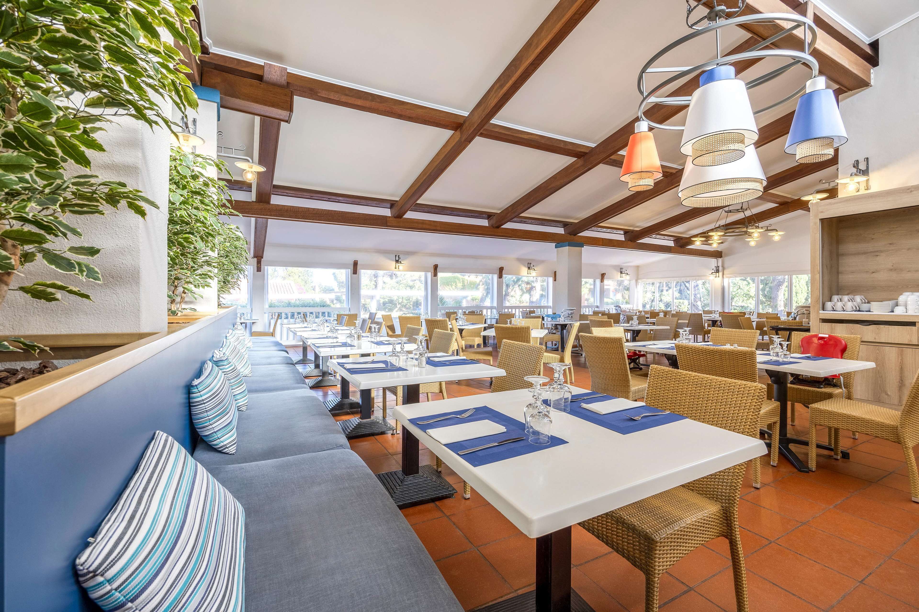 A restaurant with long tables and benches, wooden beams on the ceiling, and hanging pendant lights.