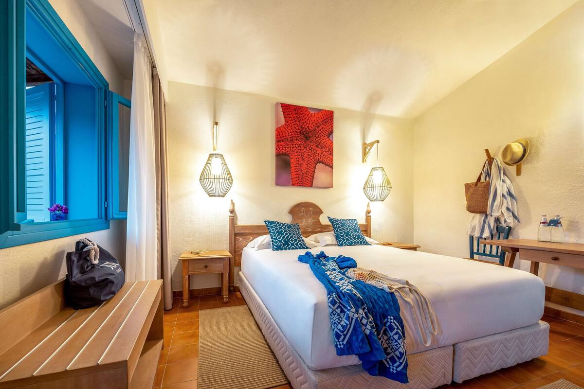 A hotel room with a large bed, two bedside tables, a bench, a desk, and a window with blue shutters.