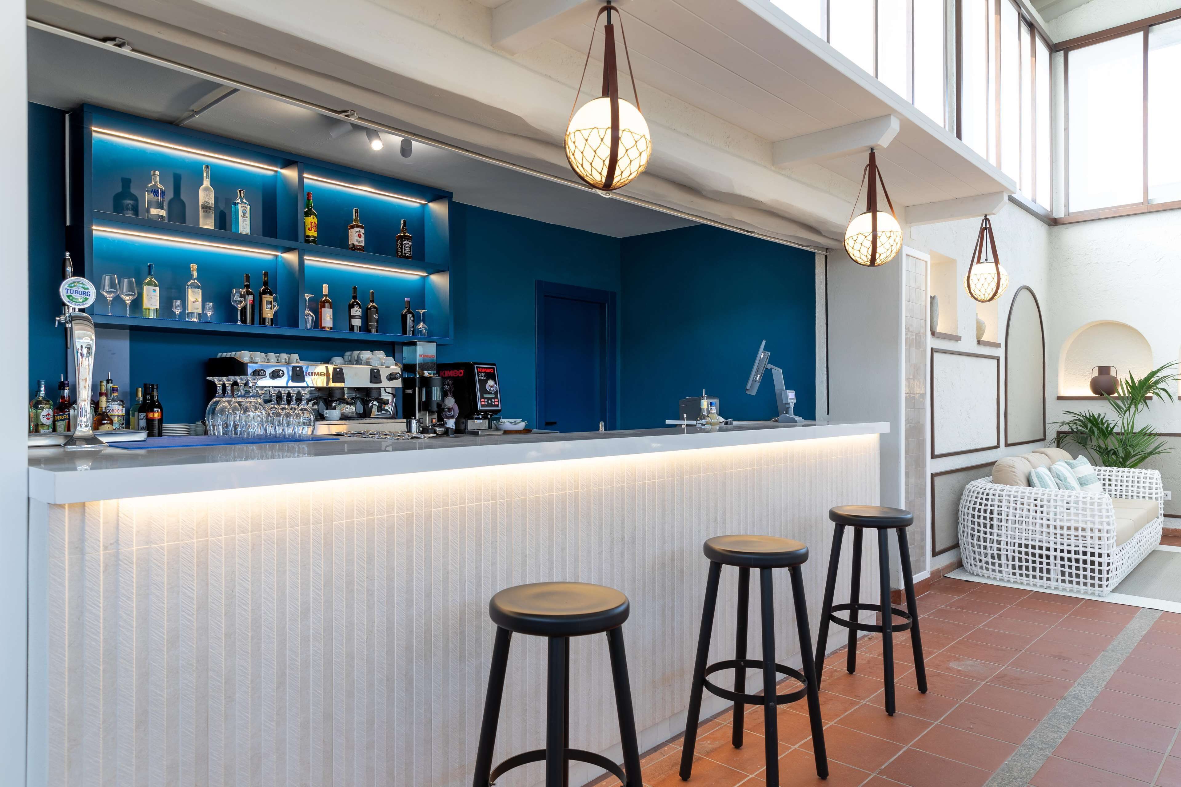 A bar area with a blue backdrop, bar stools, and a variety of bottles and glasses.
