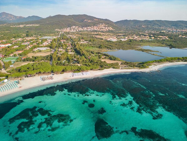 Vista aérea de un pueblo costero con playa, aguas turquesas cristalinas y colinas circundantes.