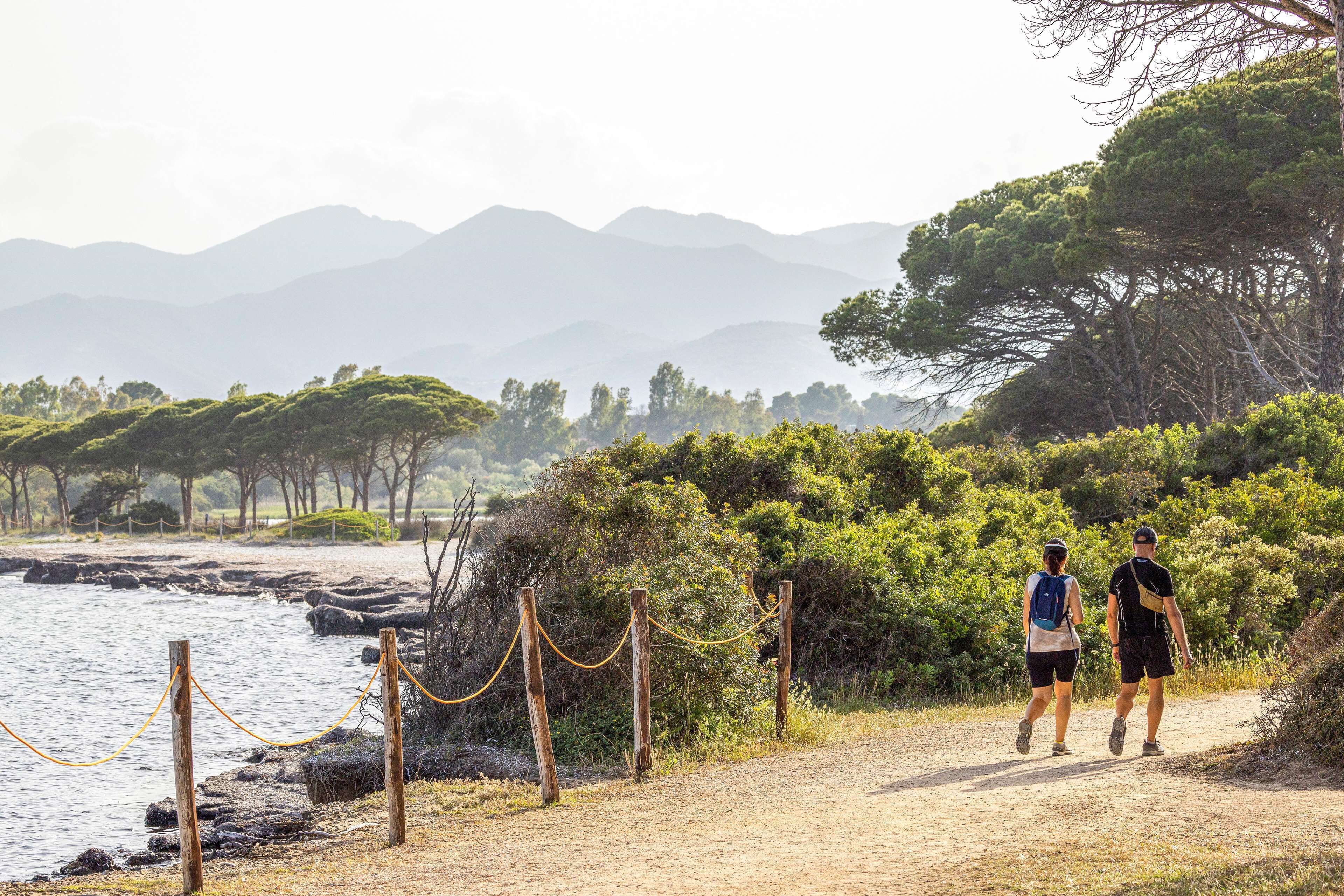 Um caminho costeiro com uma cerca de arame e estacas de madeira, que leva a um corpo d'água com montanhas ao fundo.