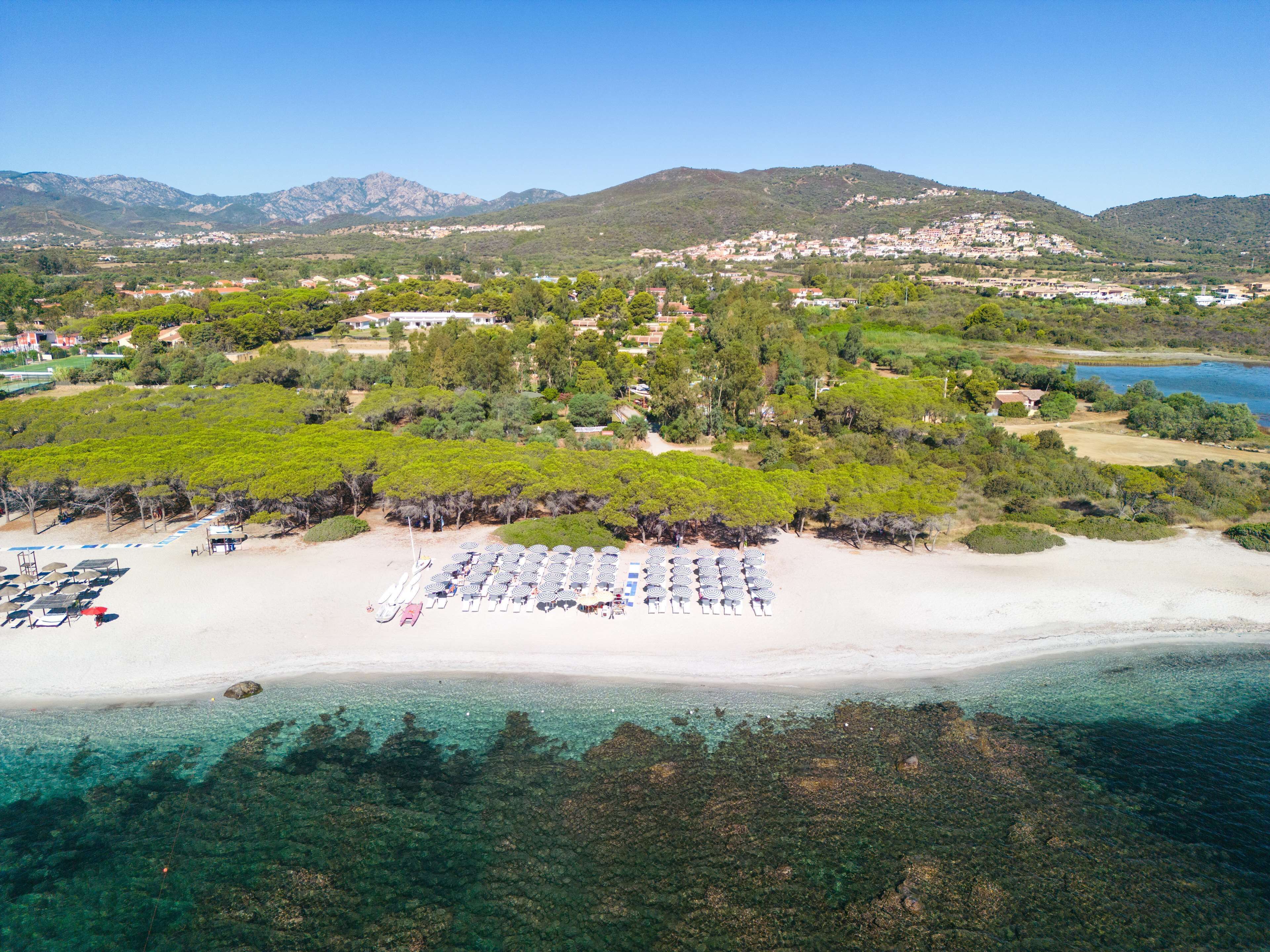 An aerial view of a beach with rows of umbrellas and lounge chairs, a clear view of the coastline, and a backdrop of hills and buildings.