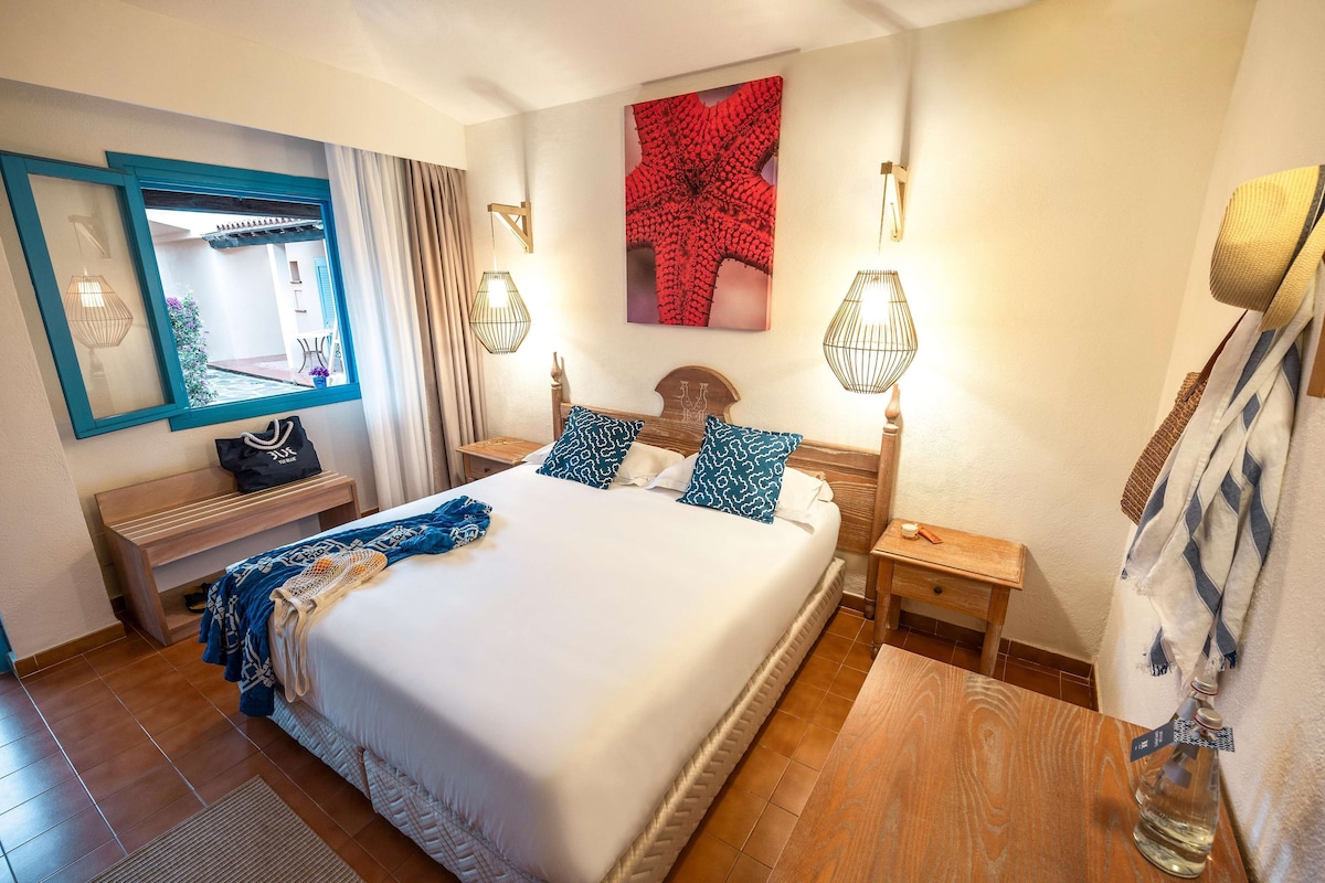 Habitación de hotel bien iluminada con una cama grande, mesitas de noche, un espejo y vistas al exterior.