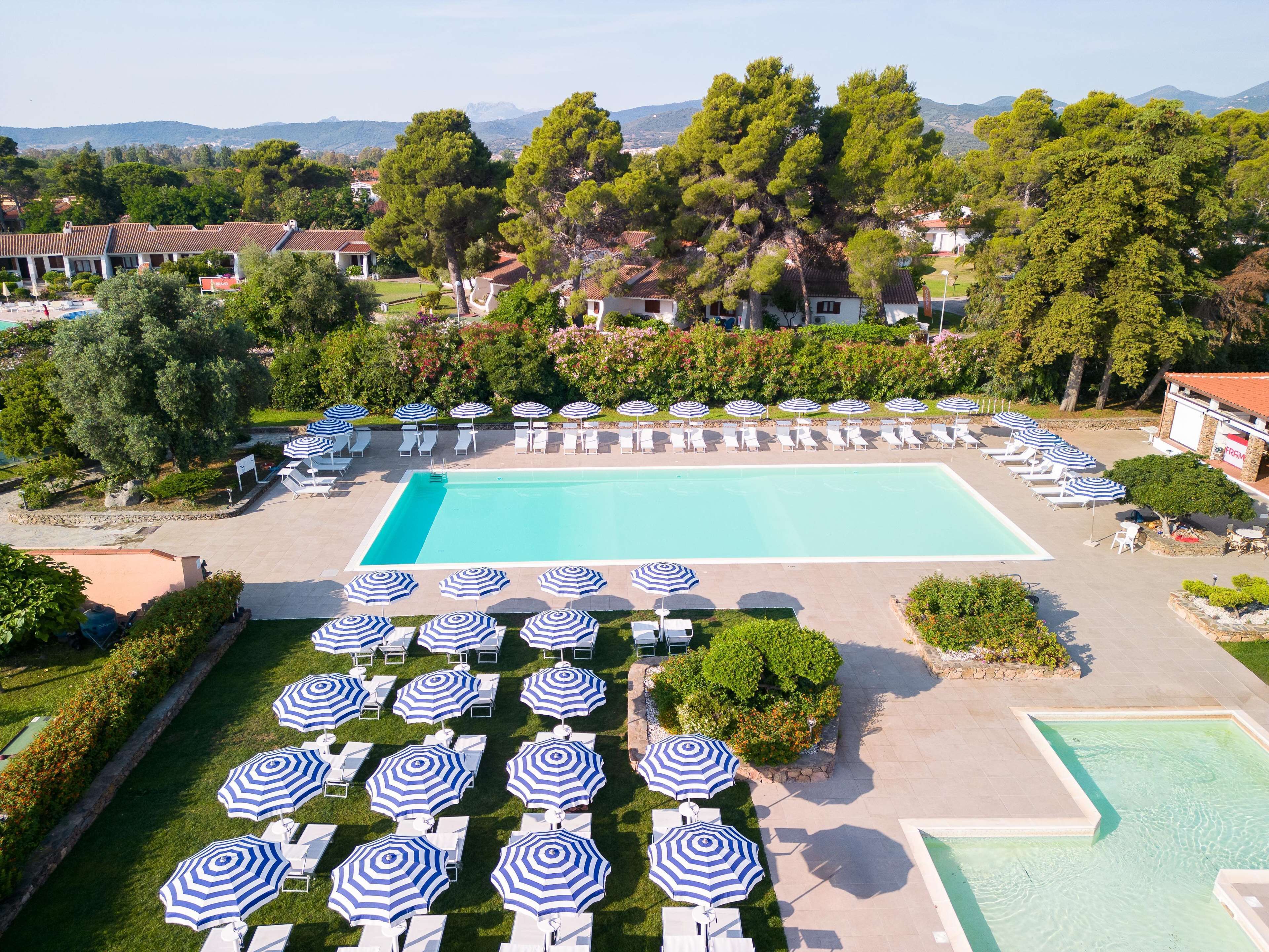 Ein großer Swimmingpool, umgeben von Liegestühlen und Sonnenschirmen, mit Blick auf Bäume und Gebäude im Hintergrund.