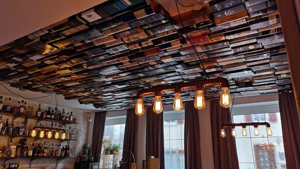 A bar area with a ceiling covered in books, a bar counter with bottles, and a window with curtains. - Hotel Restaurant Dragoner (Peiting)
