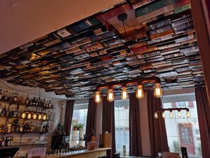 Zona de bar con techo cubierto de libros, barra con botellas y ventana con cortinas.