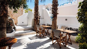 An outdoor seating area with wooden tables and chairs, surrounded by palm trees and a stone wall. - Pension George (Santorini)