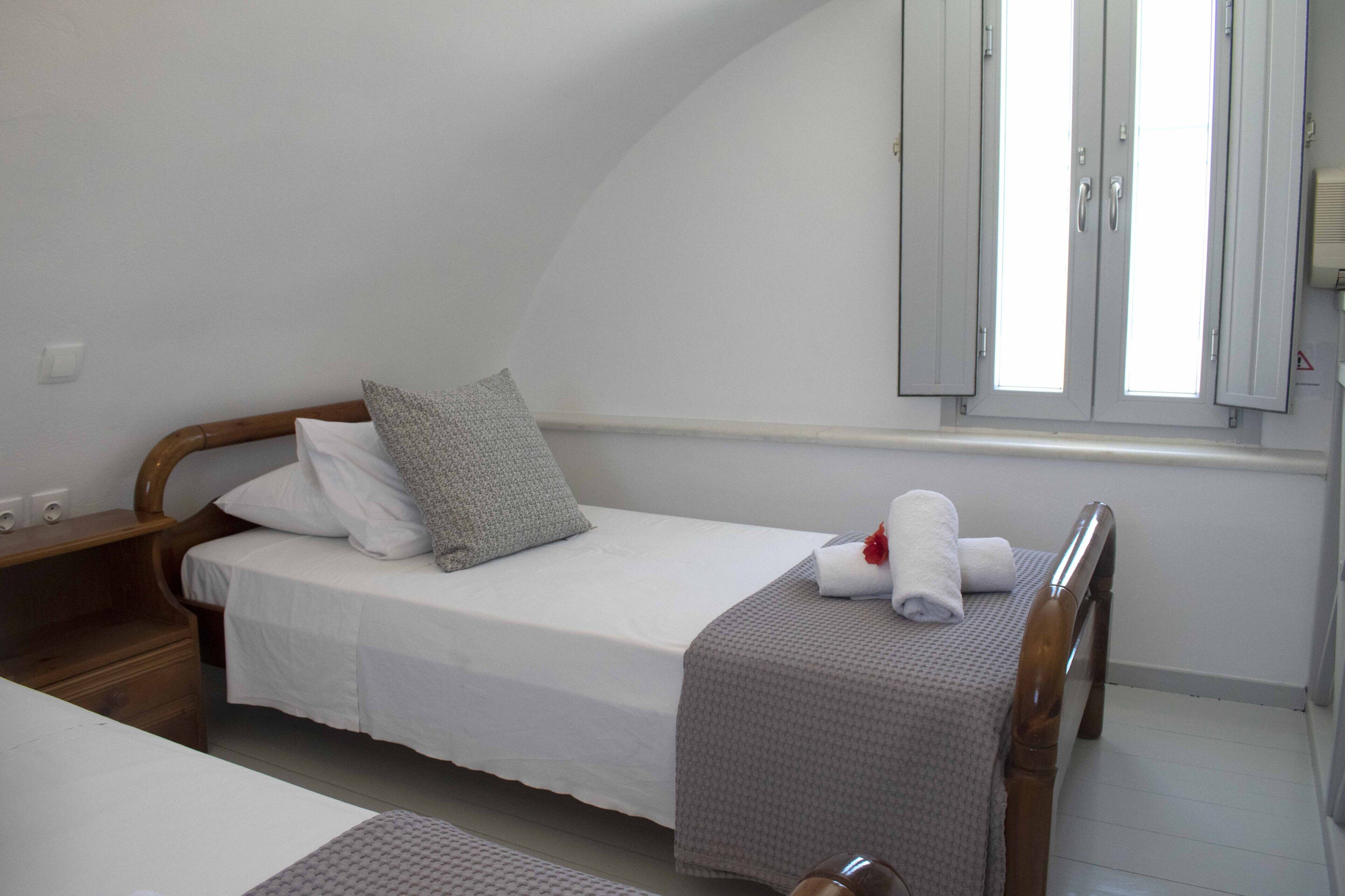A single bed with a wooden headboard, a grey pillow, and a rolled white towel on the footboard.