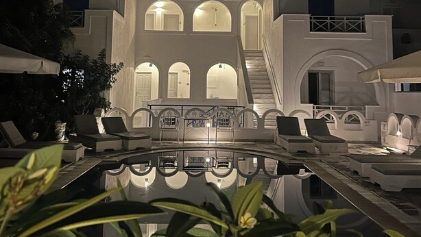 A pool area with lounge chairs and a reflection pool, surrounded by a building with arched windows and a staircase leading to an upper level. - Pension George (Santorini)
