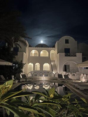 A pool area with lounge chairs and a reflection pool, surrounded by a building with arched windows and a staircase leading to an upper level. - Pension George (Santorini)
