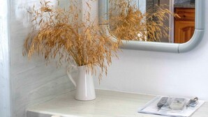A white table with a vase of dried plants, a chair, and a mirror reflecting a kitchen area. - Pension George (Santorini)