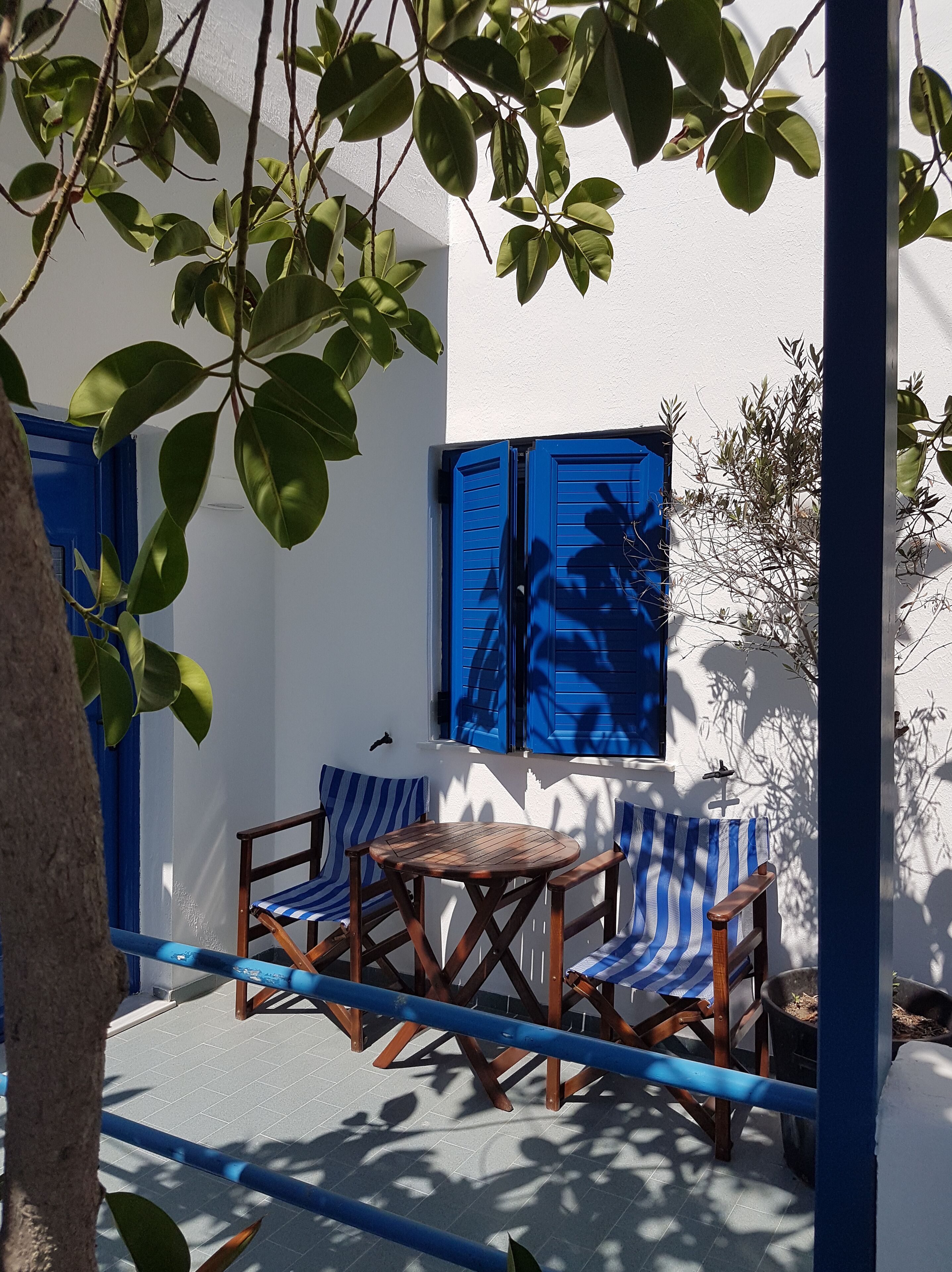 Ein kleiner Außenbereich mit Tisch und zwei Stühlen, einer blauen Tür und einem Fenster mit blauen Rollläden.