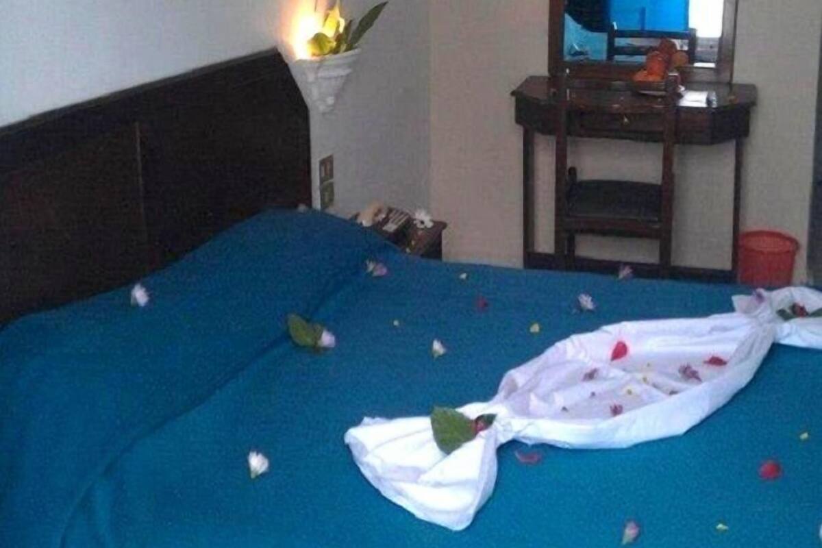 Una cama con sábanas azules y pétalos de rosa esparcidos, una mesita de noche con un espejo y una planta pequeña en la mesita.