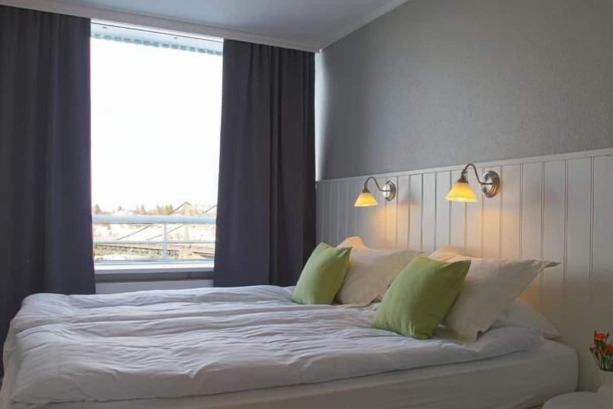 Habitación de hotel con una cama grande, dos lámparas fijadas a la pared, un ventanal con cortinas y vista a un puente.