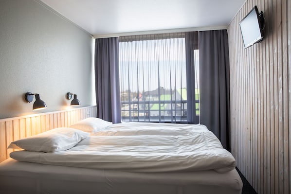 A hotel room with a bed, two wall-mounted lamps, a TV, and a window with curtains.