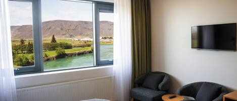 A hotel room with a large window offering a view of a river and mountains, a bed, two armchairs, a small table, and a flat-screen TV mounted on the wall.