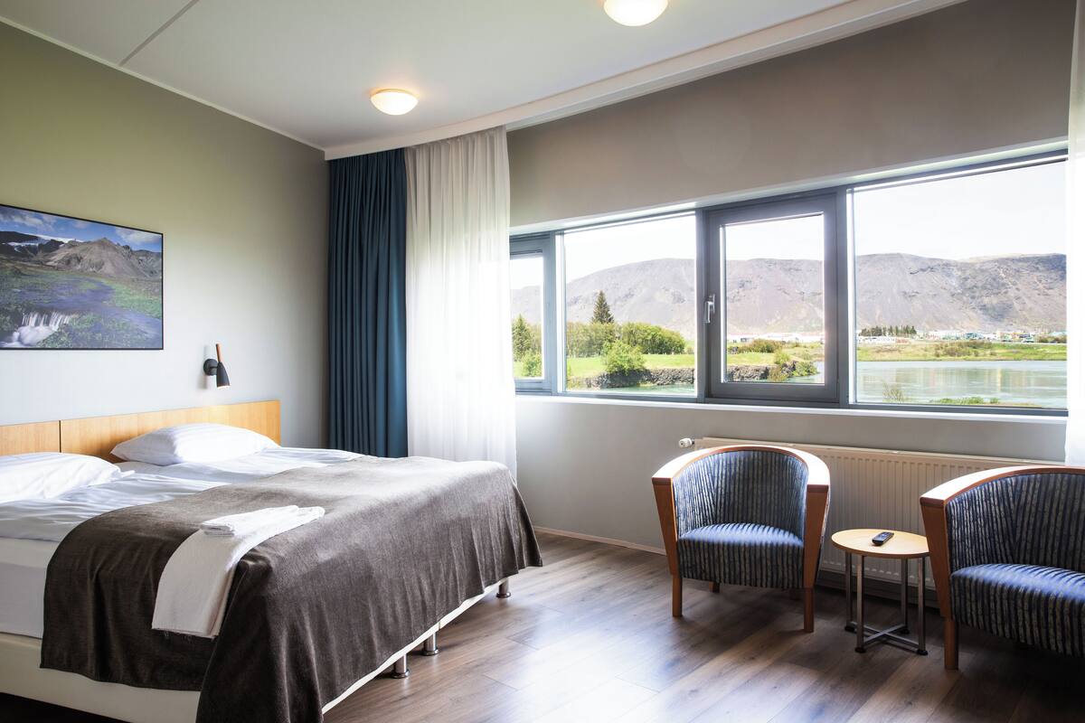 Habitación de hotel con una cama grande, dos sillones, una mesita y vistas a montañas y un lago a través de la ventana.