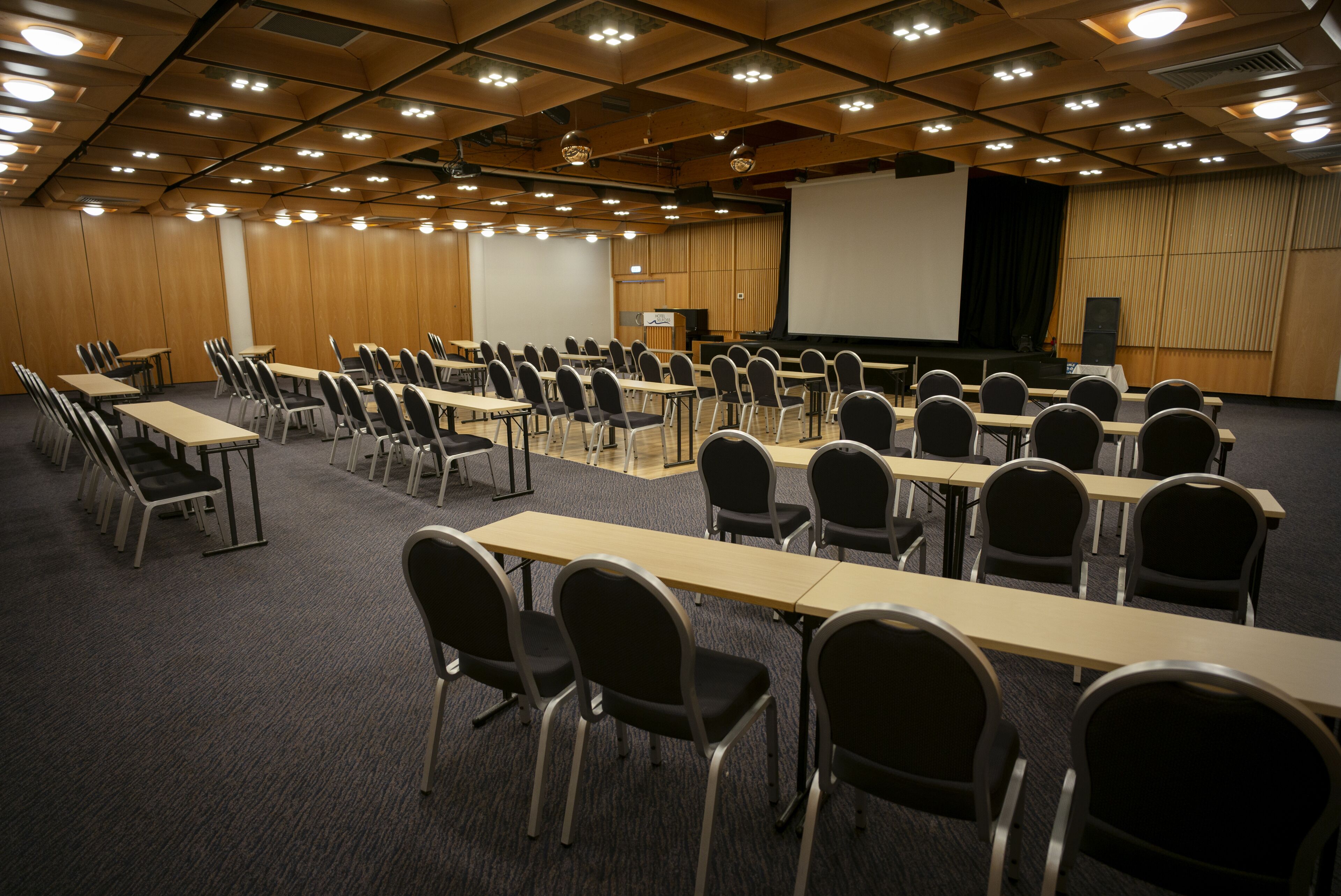 Une salle de conférence avec des rangées de tables et de chaises, un écran de projection et une scène.