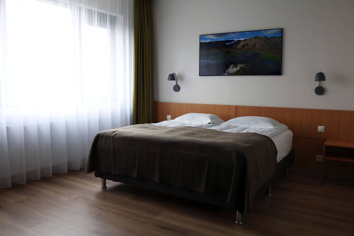 Habitación de hotel con una cama grande, dos mesitas de noche, una mesita auxiliar, un cuadro en la pared y cortinas translúcidas en la ventana.