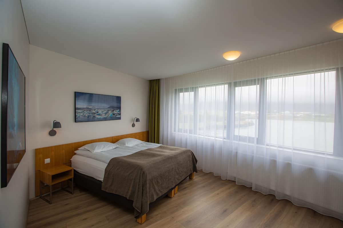 Habitación de hotel con cama, mesita de noche, lámpara de pared, un cuadro y un gran ventanal con cortinas translúcidas.