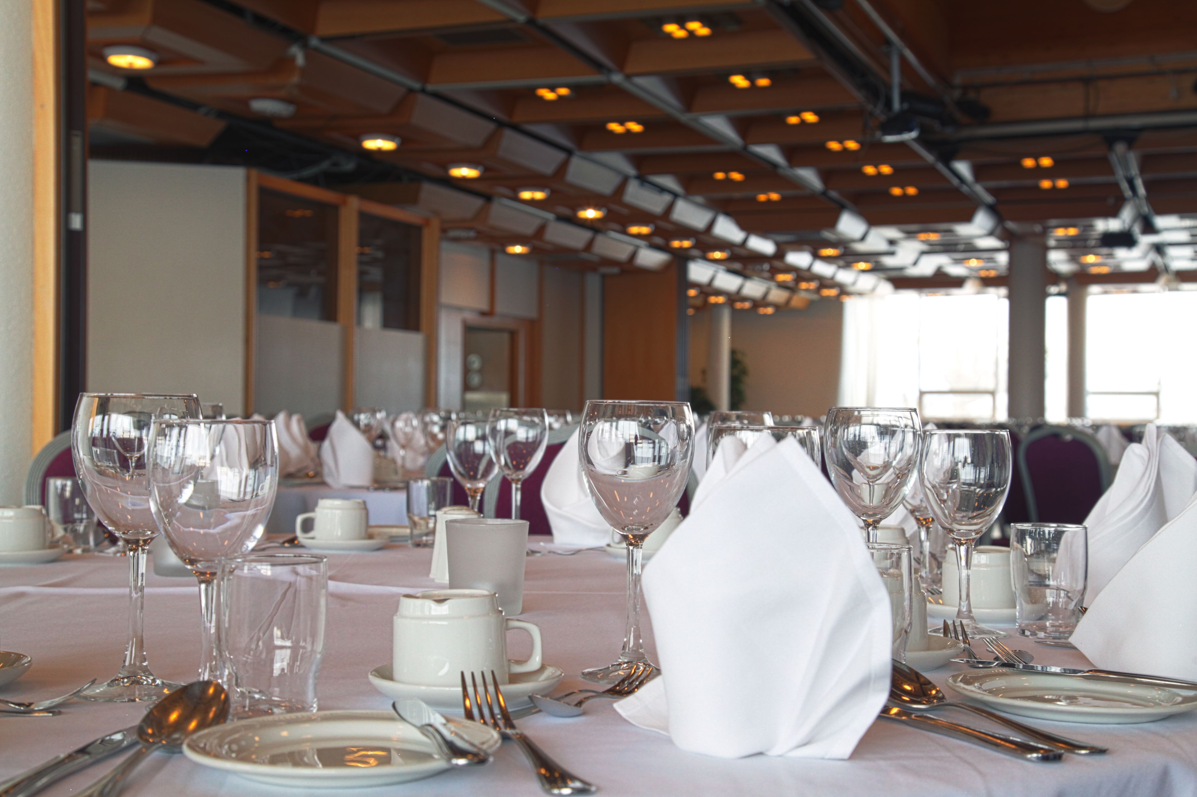 Un salón de banquetes con mesas dispuestas para un evento formal, con manteles blancos, cristalería transparente y cubertería de plata.