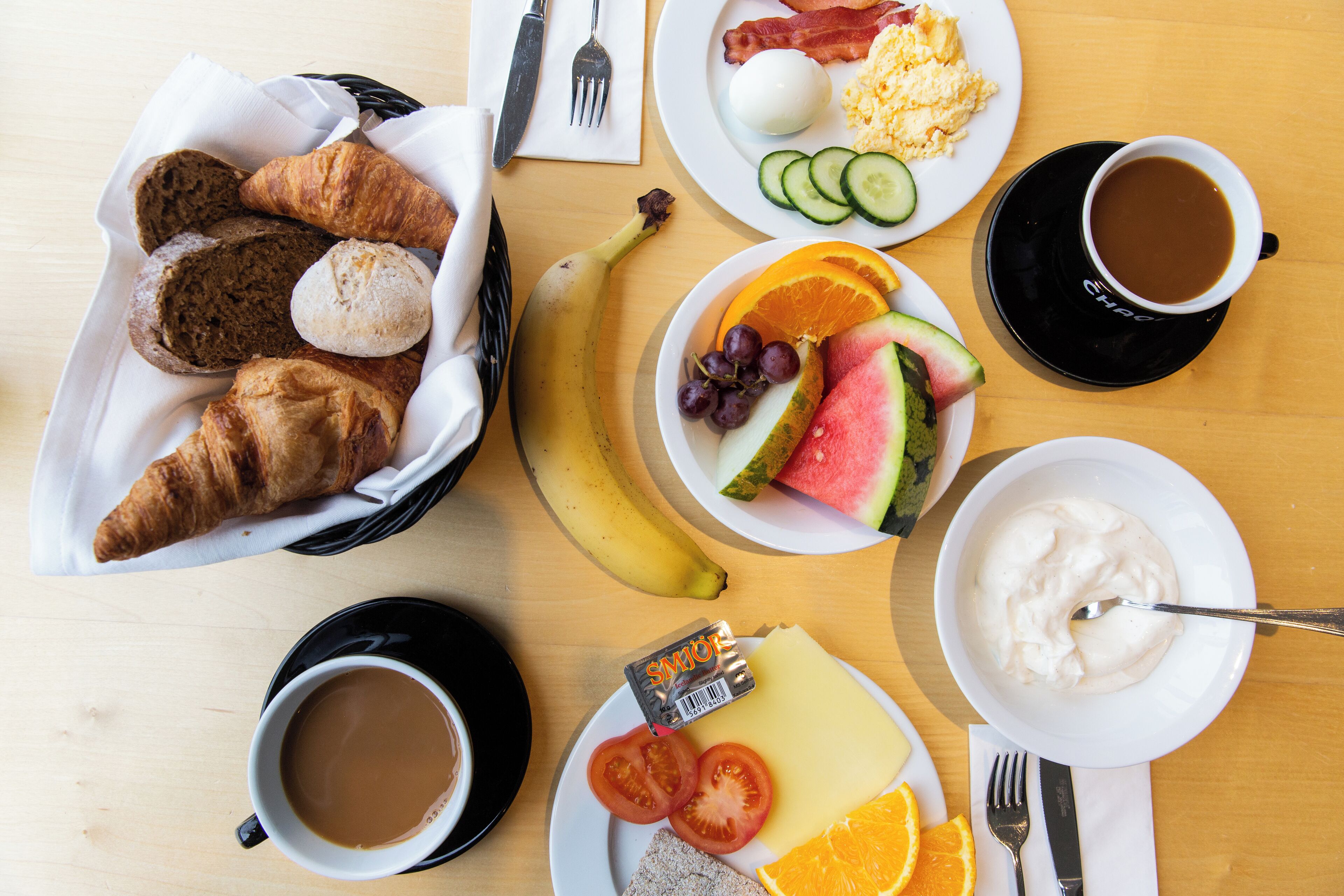 A breakfast table with croissants, eggs, bacon, a banana, tomatoes, grapes, watermelon, and coffee.