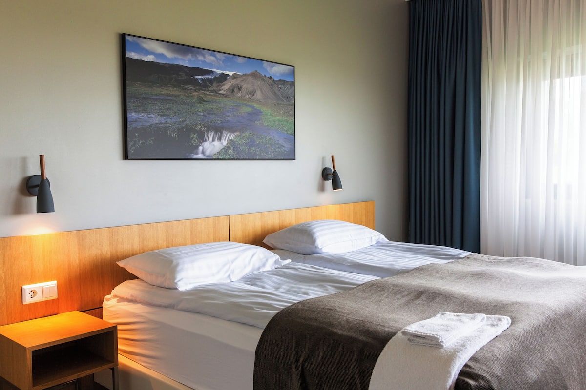 Una habitación de hotel con cabecera de madera, una cama con ropa blanca, una mesita de noche con lámpara, un cuadro de un paisaje montañoso y una ventana con cortinas traslúcidas.