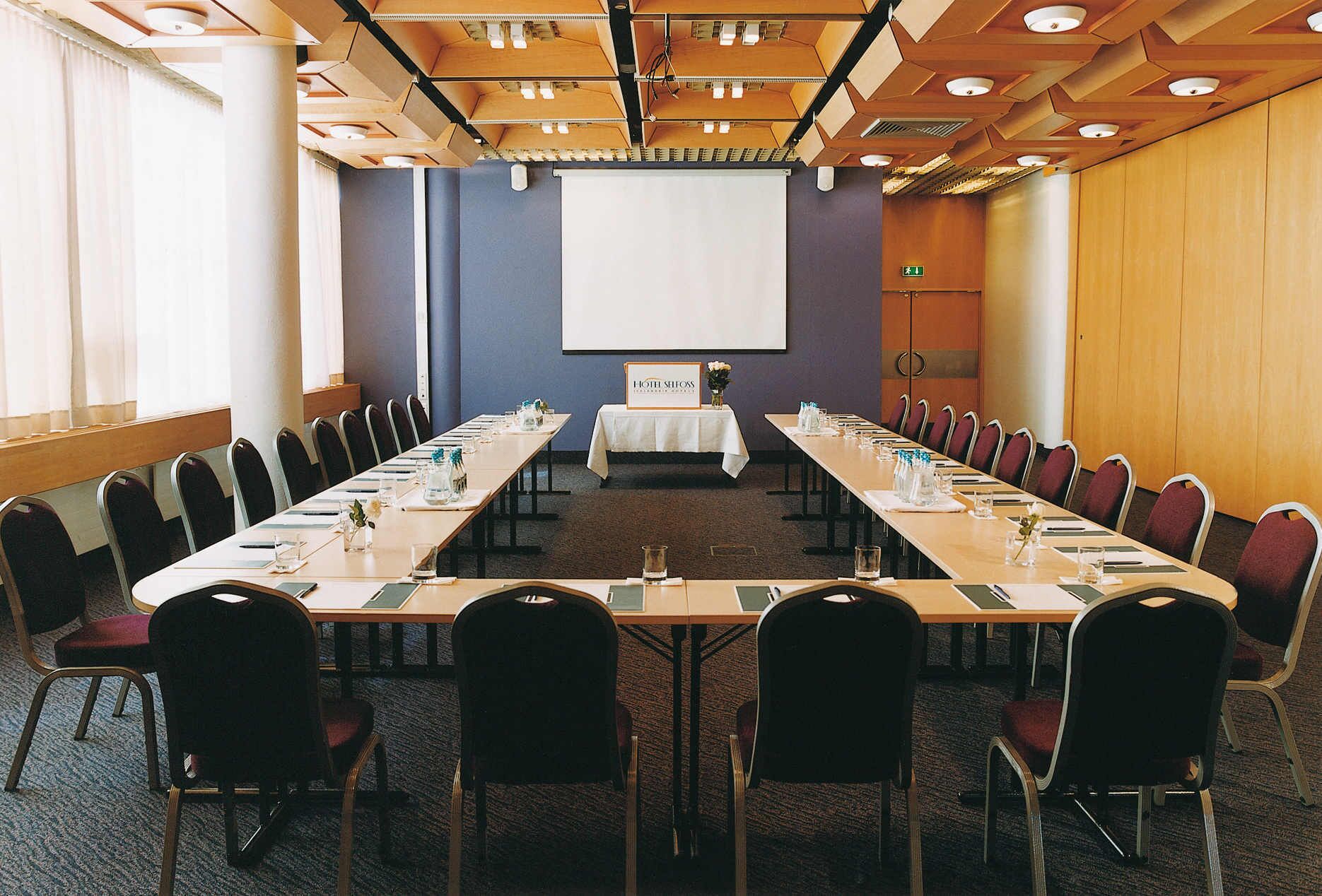 Un salón de conferencias con una larga mesa, sillas, una pantalla de proyector y un letrero con el nombre del hotel.