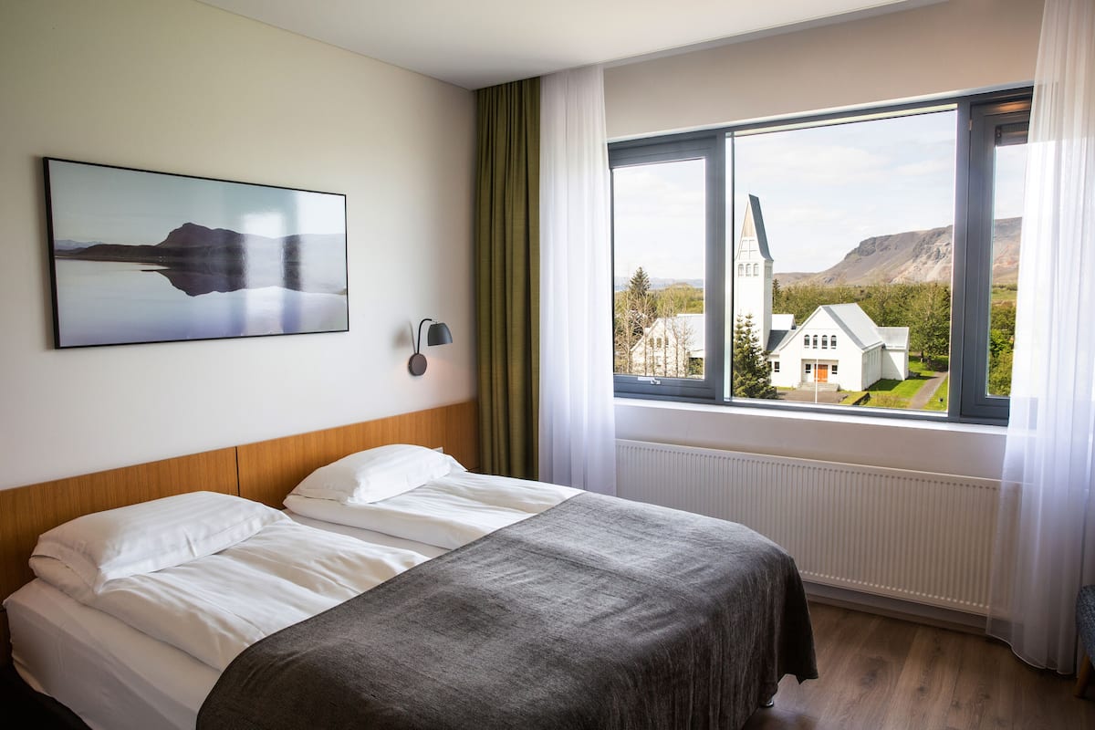 Habitación de hotel con una cama, un cuadro de un paisaje, una ventana con vista a una casa y montañas, y una lámpara en la pared.