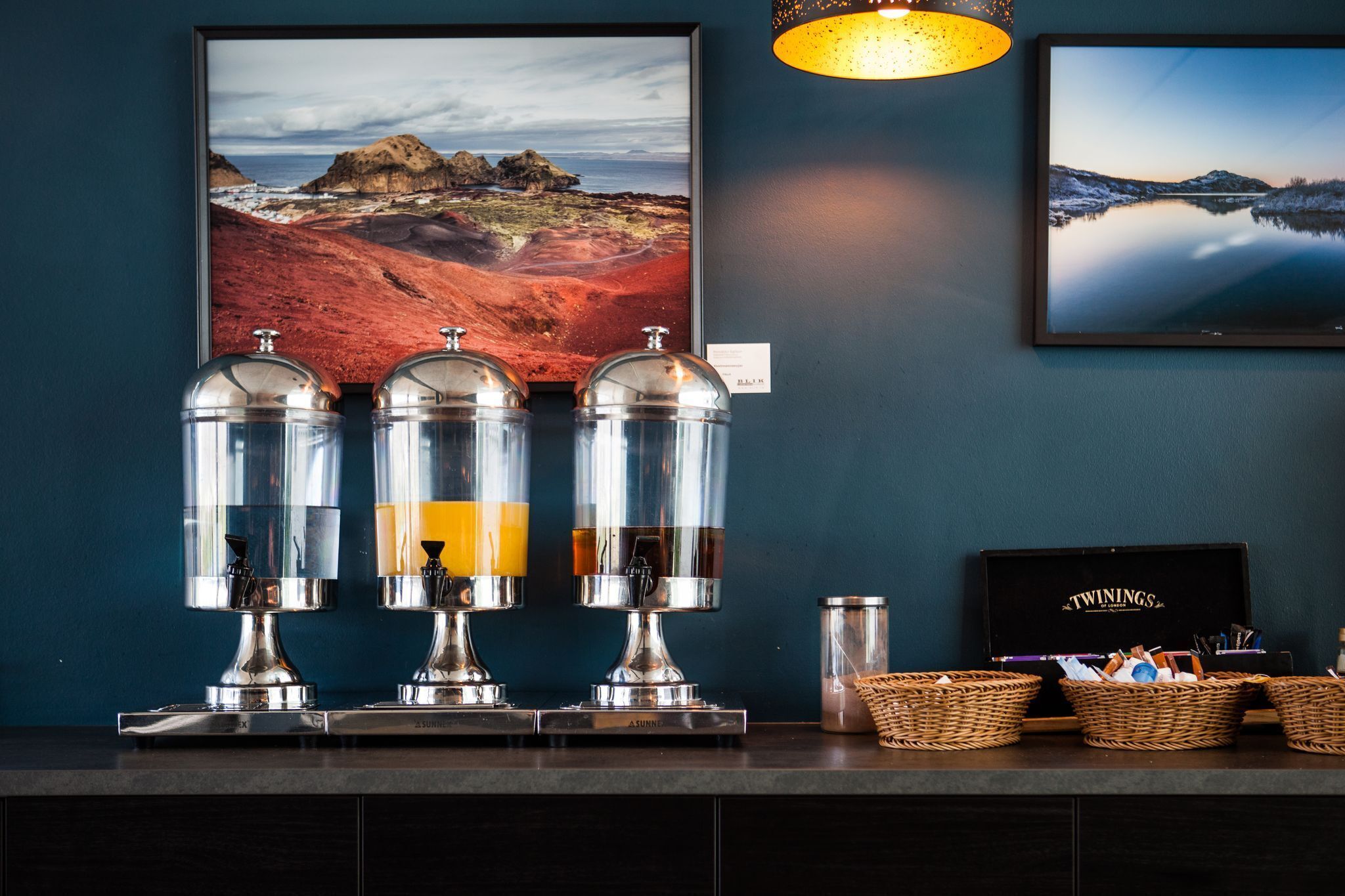 A bar area with three beverage dispensers, two framed landscape pictures, and a basket of snacks.