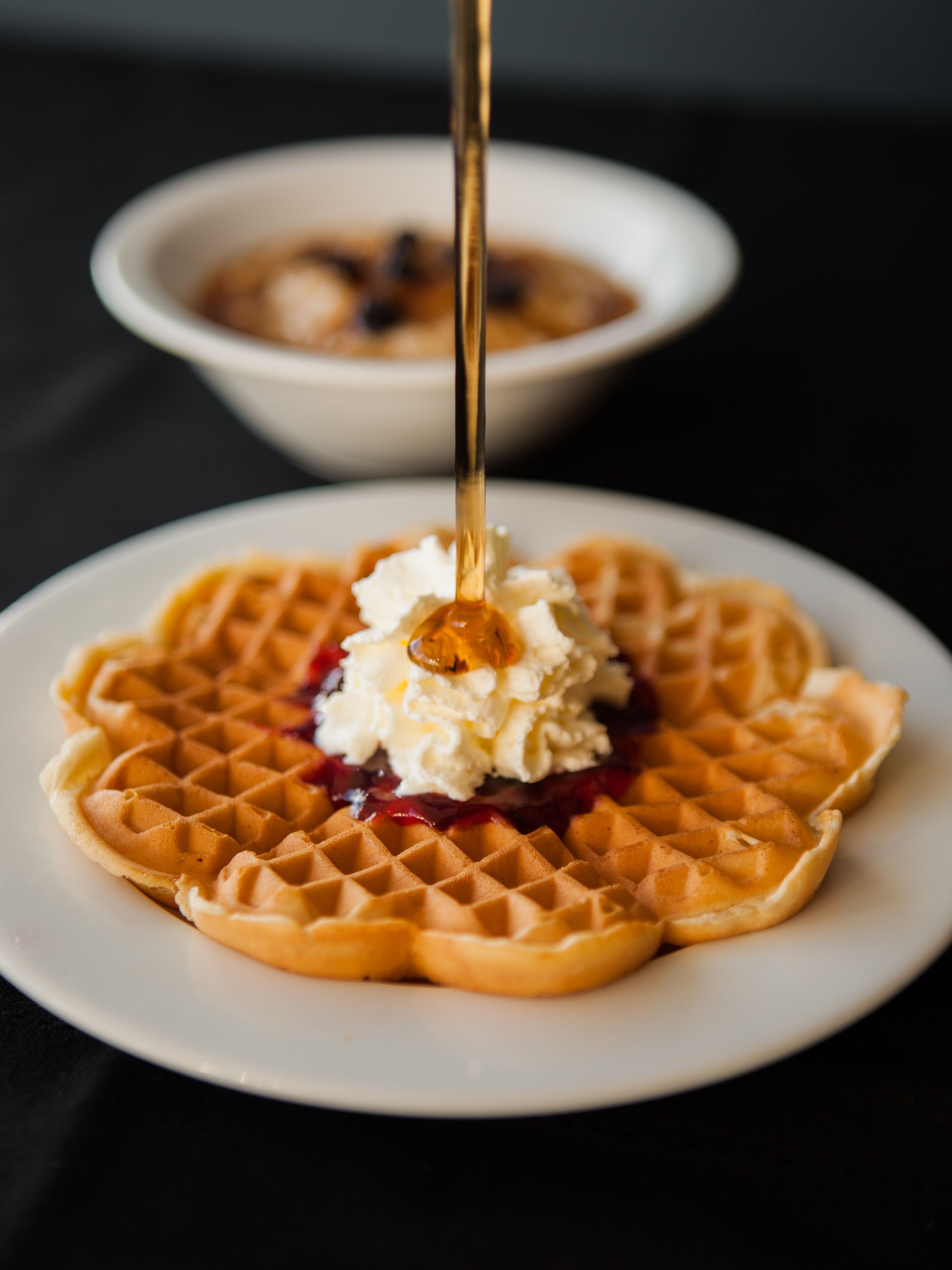 A close-up of a waffle with whipped cream and syrup, on a white plate.