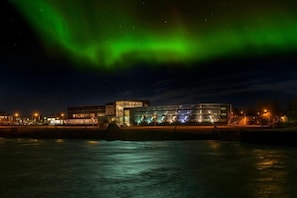 A modern building with a glass facade by a waterfront, reflecting lights and the green aurora.