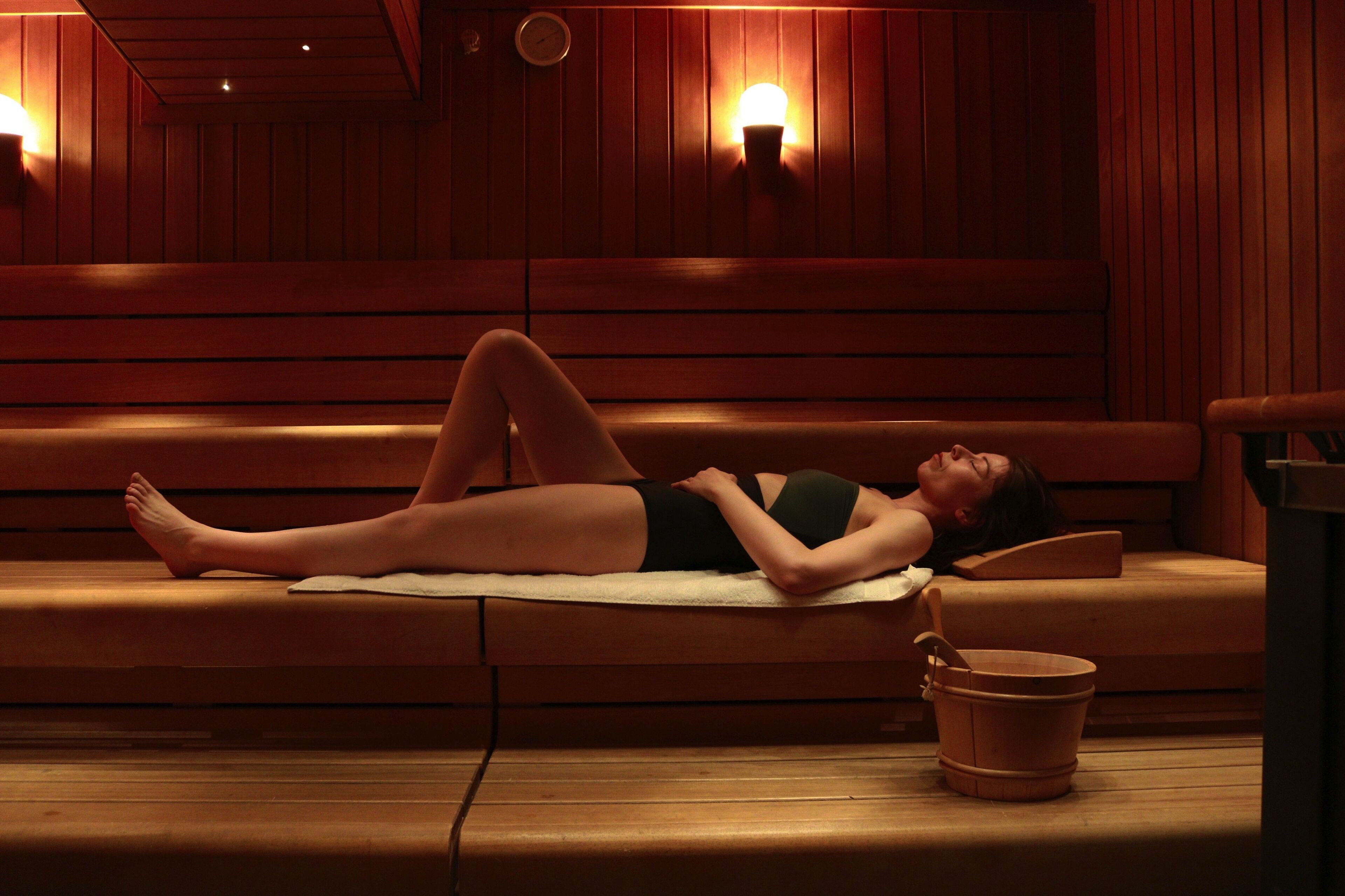 A person lying on a wooden bench in a dimly lit room with a wooden floor and a bucket on the floor.