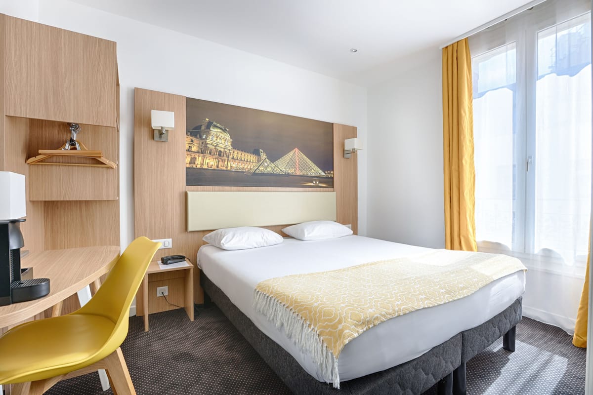 A hotel room with a bed, a desk, a chair, a painting of a bridge, and a window with curtains.