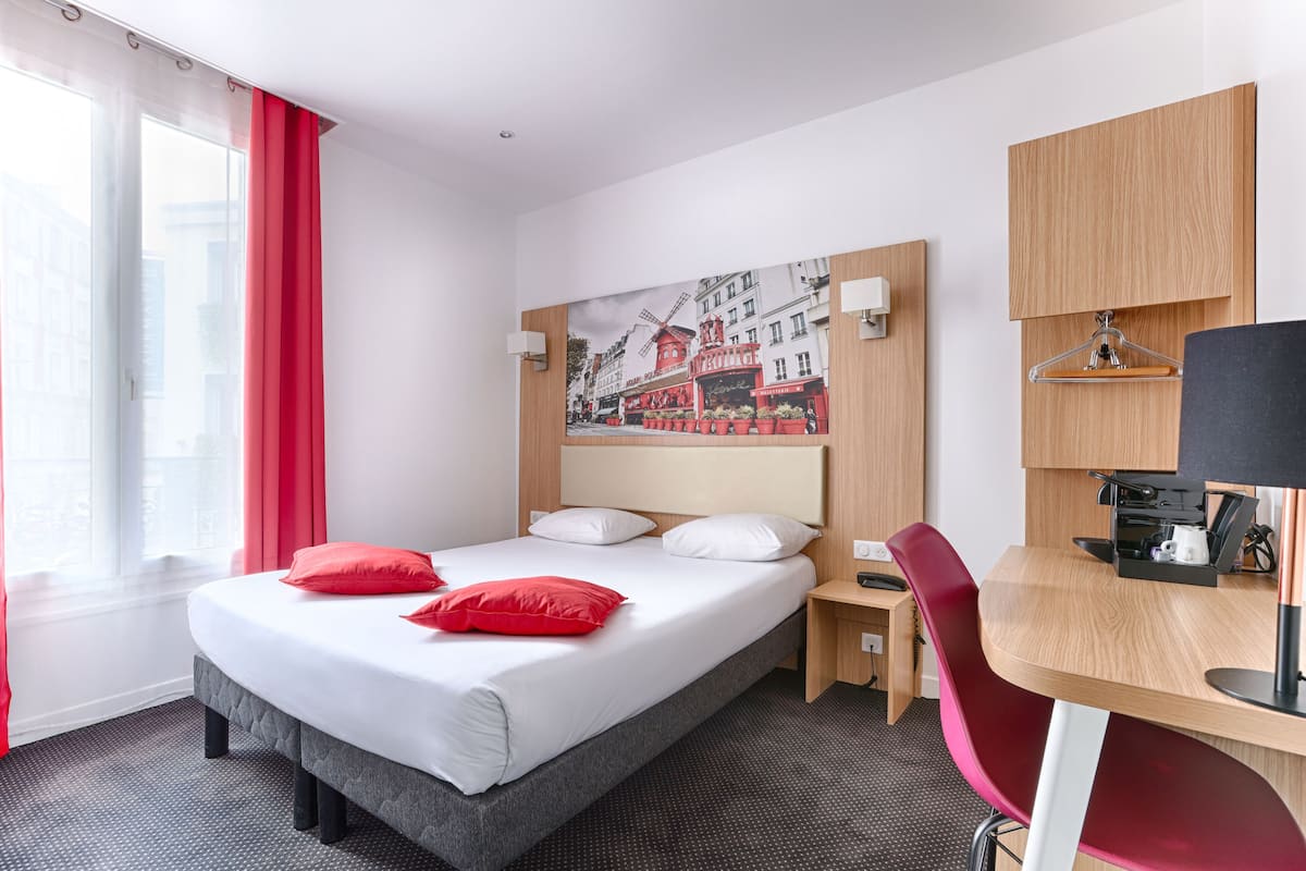 A hotel room with a bed, a desk, a chair, a painting on the wall, and a window with curtains.