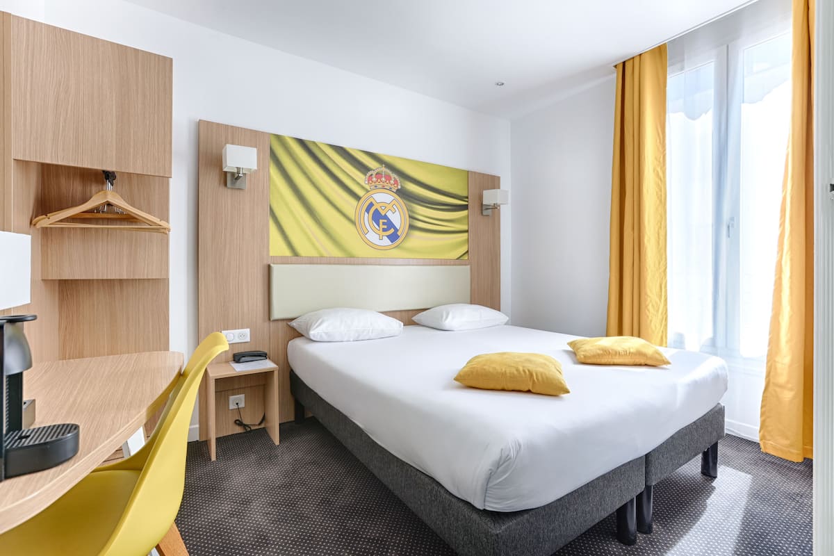 A hotel room with a bed, a desk, a chair, a coat rack, and a wall-mounted light.