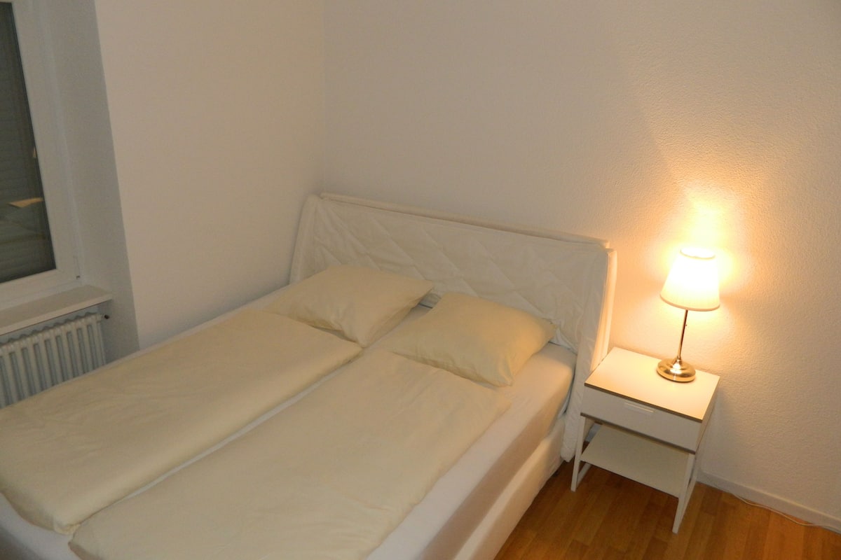 Una cama individual con cabecera blanca y dos almohadas, una mesita de noche con lámpara, y una ventana con persianas.