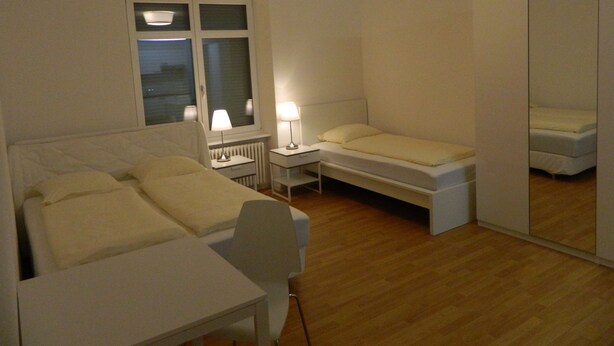 A bedroom with a bed, bedside tables, a chair, a desk, a mirror, and a window with blinds. - Swiss Star Delsbergerallee (Basel)