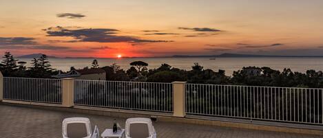 Une terrasse avec deux chaises blanches et une table, d’où l’on peut admirer le coucher de soleil sur la mer.