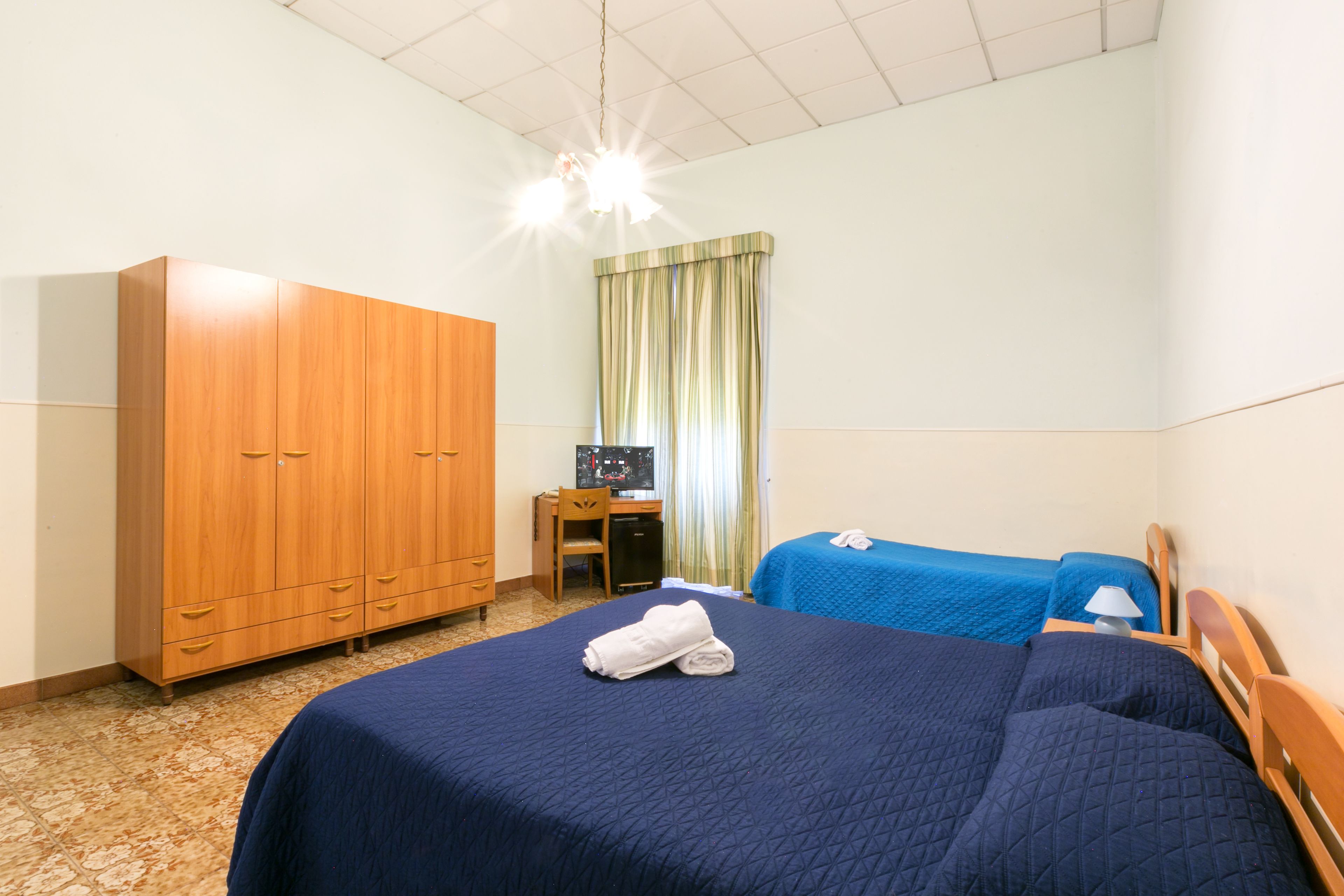 A hotel room with a large wardrobe, a desk, two beds with blue bedding, and a window with curtains.