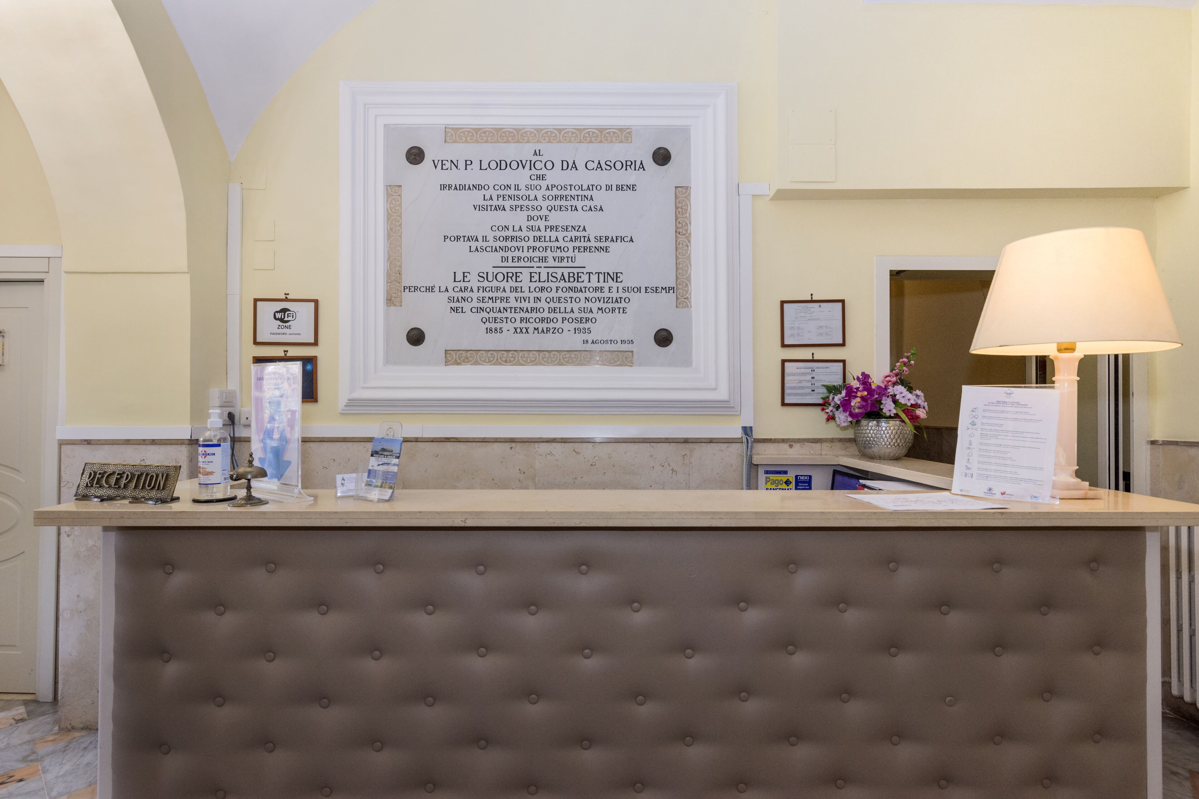 A reception desk with a framed plaque, a reception sign, a lamp, and a vase with flowers.