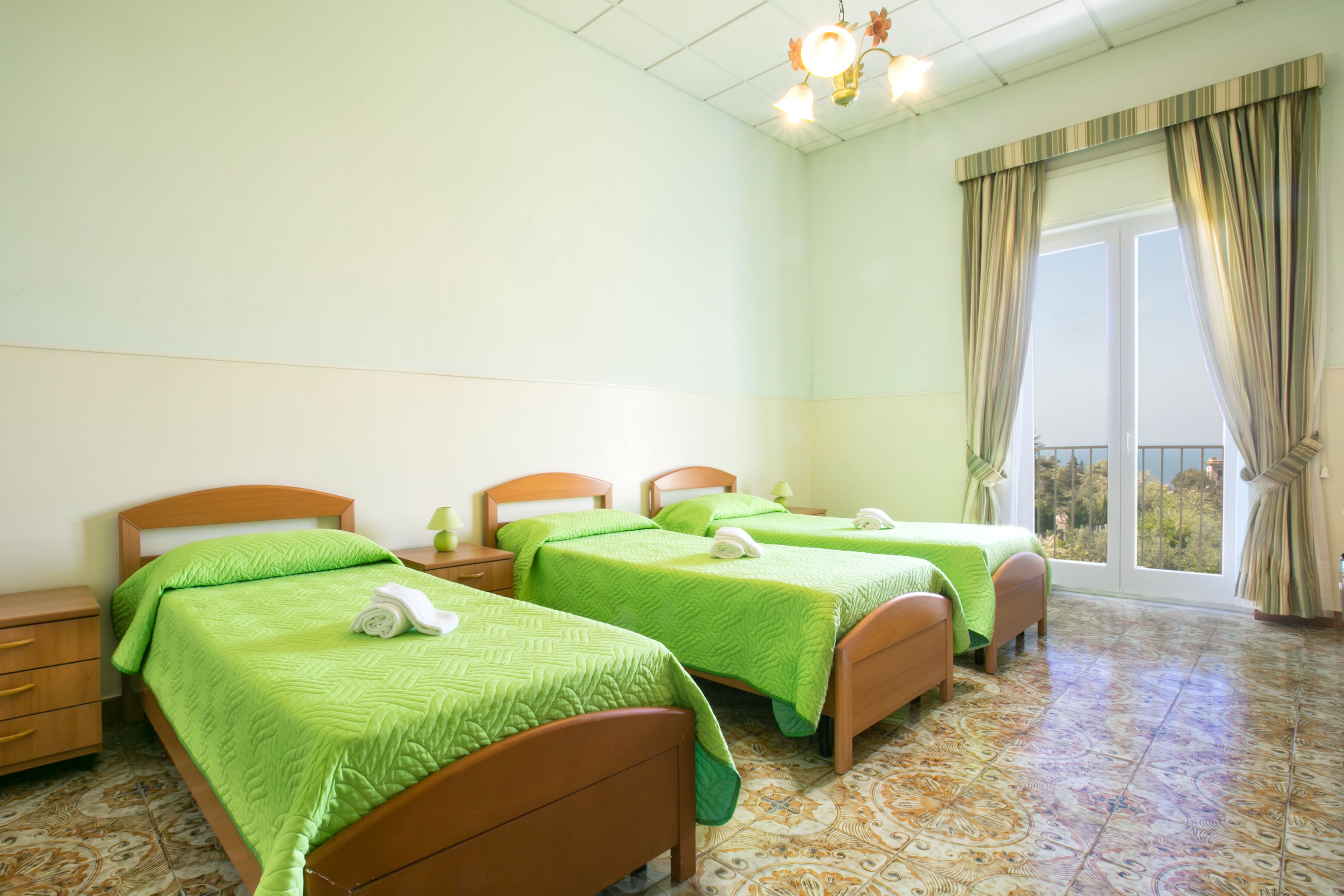 Une chambre d’hôtel avec trois lits simples, une grande fenêtre avec des rideaux, un sol à motifs et un luminaire au plafond.